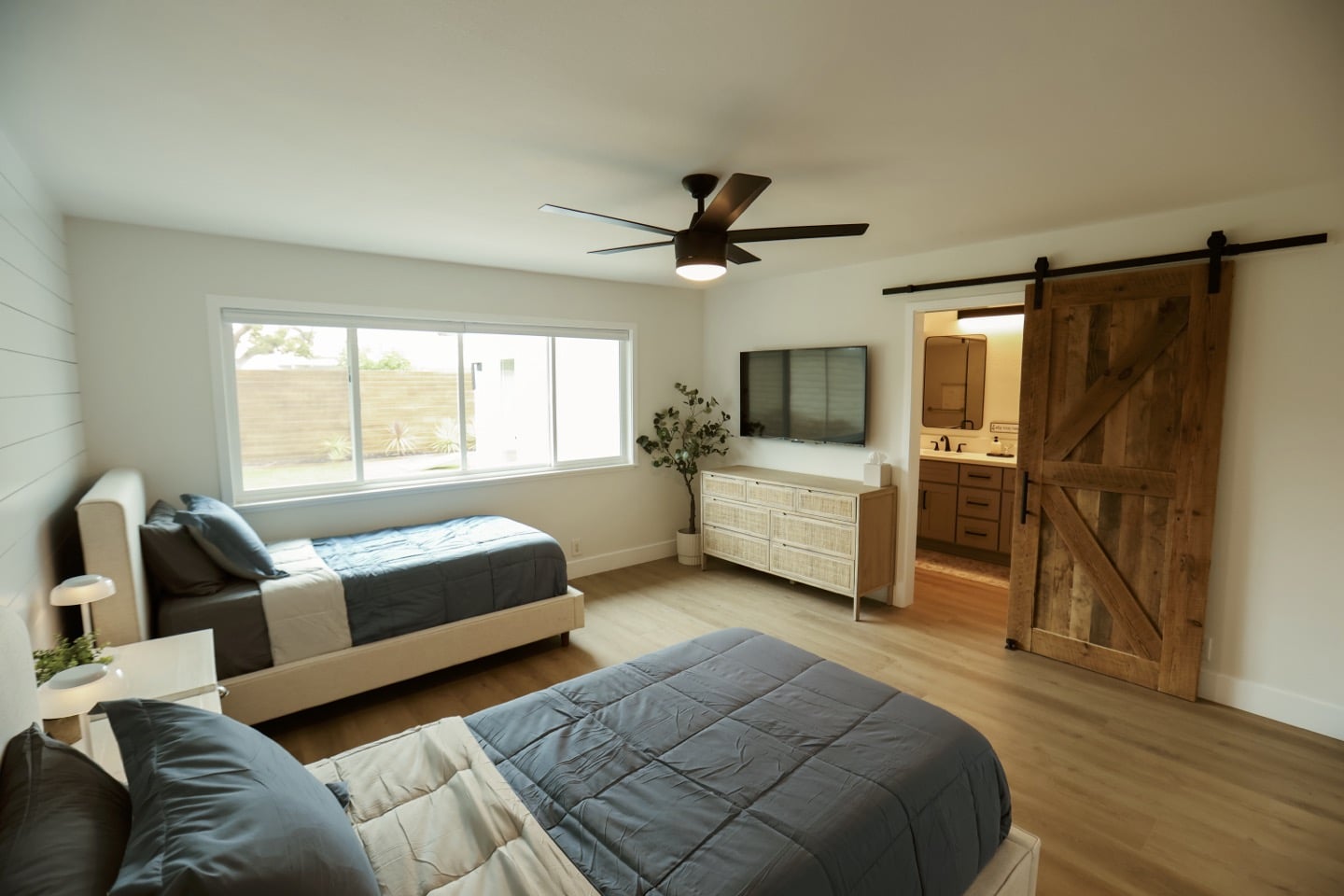 CariHouse1JaredSislinPhoto-21-copyjpg - Lonestar Mental Health Bedroom interior with two beds, a sliding barn door to a bathroom, and a modern ceiling fan.