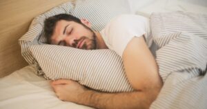 Man sleeping in bed, cuddling a striped pillow under a light duvet.
