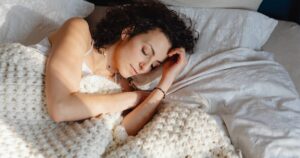 Woman sleeping on her side in bed, wrapped in a thick cream knit blanket.