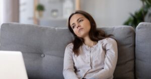 Woman sitting on a gray sofa with a laptop nearby, looking tired and lost in thought in a home setting.