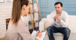 Therapist takes notes on a clipboard while a man talks in a living room–style therapy session in the background.