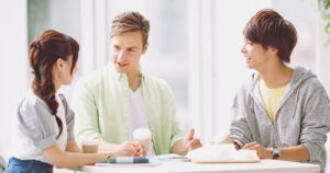 Three young friends sit at a bright table chatting with takeaway coffees in a cafe-like setting.