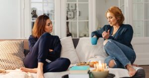 Two women sit on a bright living room sofa, chatting; one holds a blue mug while gesturing, with cookies and candles on a nearby table.