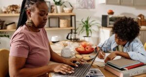 Two people do homework at a kitchen table: one types on a laptop while the other writes in a notebook, with books, apples, and cookies nearby.