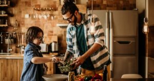 Father and son chopping greens to prepare a salad in a warm, wood-toned kitchen.