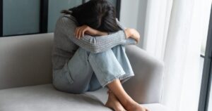 Young woman sits on a gray couch with her head down and arms wrapped around her knees, conveying distress or sadness.