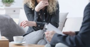 Woman with curly hair sits on a sofa, resting her chin as a counselor reviews papers across from her; a cup sits on the table nearby.