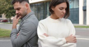 Two adults stand back-to-back with arms crossed, looking down, outdoors near a modern building.