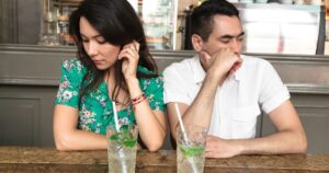 Woman in a green floral dress and man in a white shirt sit at a wooden bar, both looking away with somber expressions; two minty drinks with straws in front.