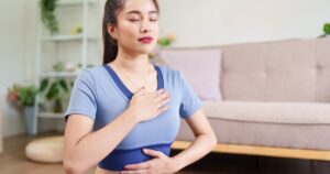 Woman in a light blue athletic top sits indoors with eyes closed, hand over her chest and other hand on her abdomen for diaphragmatic breathing in a living room setting.