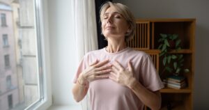 Woman standing by a window with eyes closed, hands on her chest, practicing deep breathing.