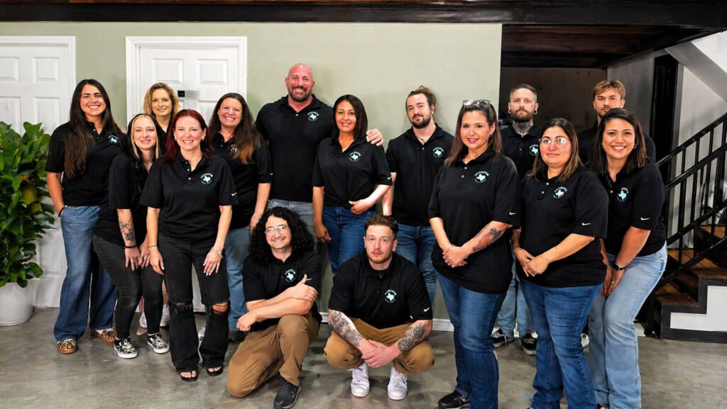 Group of 15 employees in matching black polo shirts with a logo, posing together indoors against a light green wall.