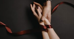 Two hands gently hold an injured or bound forearm wrapped with a red ribbon against a dark background, suggesting restraint or care.