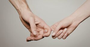 Two people link pinkies in a promise, hands touching against a neutral background.