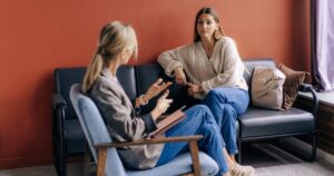 Counselor and client in a therapy room: the counselor sits in a chair with a notebook, talking as the client on the sofa listens.