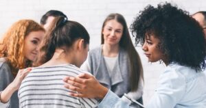 A diverse group of friends offering emotional support to a woman facing away from the camera in a bright room.