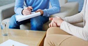 Therapist writing on a clipboard during a counseling session as a client sits with hands folded on a coffee table