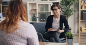 - Lonestar Mental Health Professional woman with short hair and glasses sits in a blue armchair, taking notes on a clipboard during a consultation in a bright, modern office.
