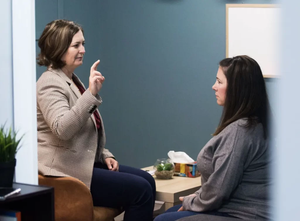 Two women in a small office during a counseling session; one speaks with a raised finger in emphasis.